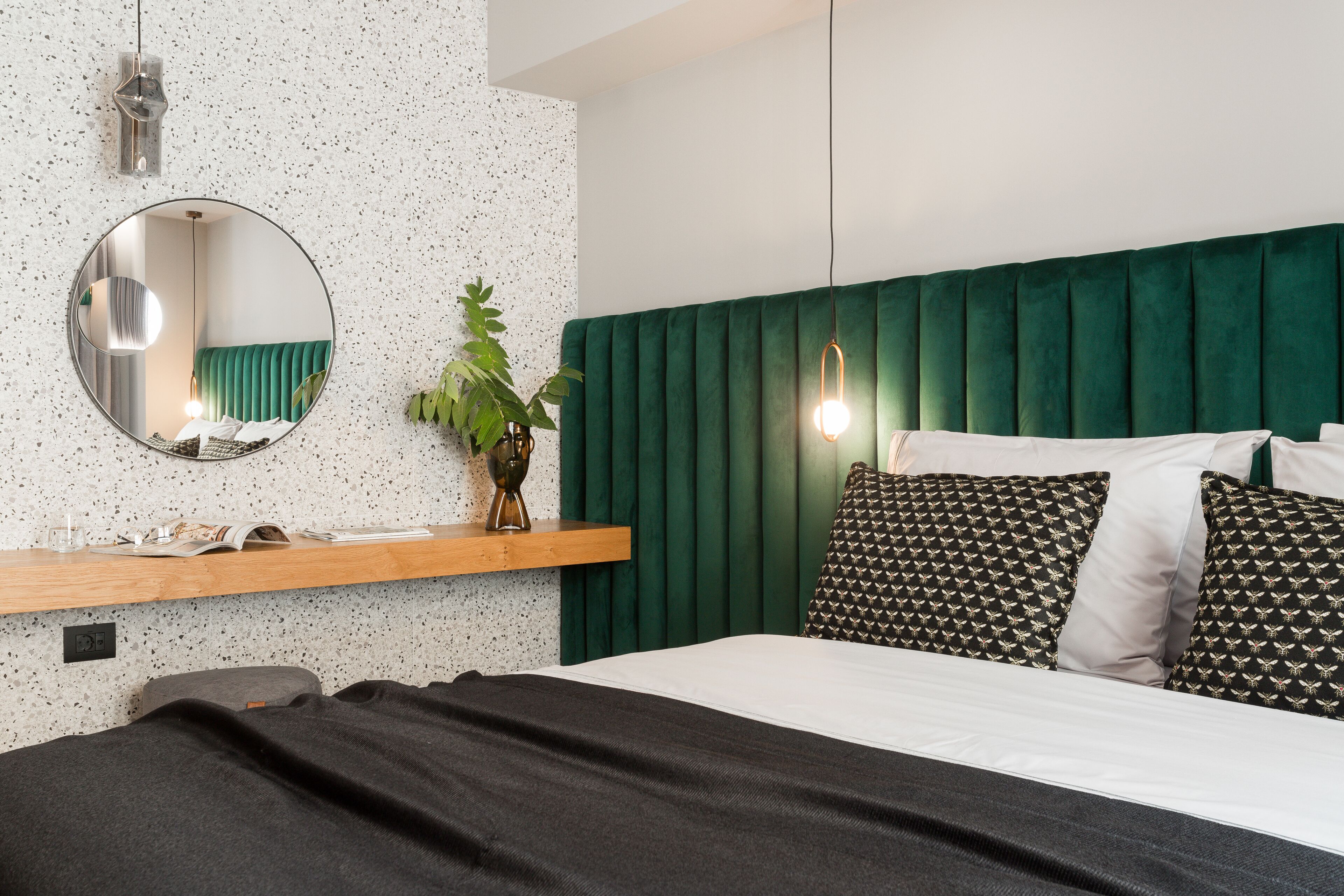 A bedroom with a green upholstered headboard, a round mirror, a wooden shelf with a plant, and a bed with patterned pillows.