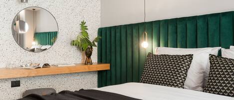 A bedroom with a green upholstered headboard, a round mirror, a wooden shelf with a plant, and a bed with patterned pillows.