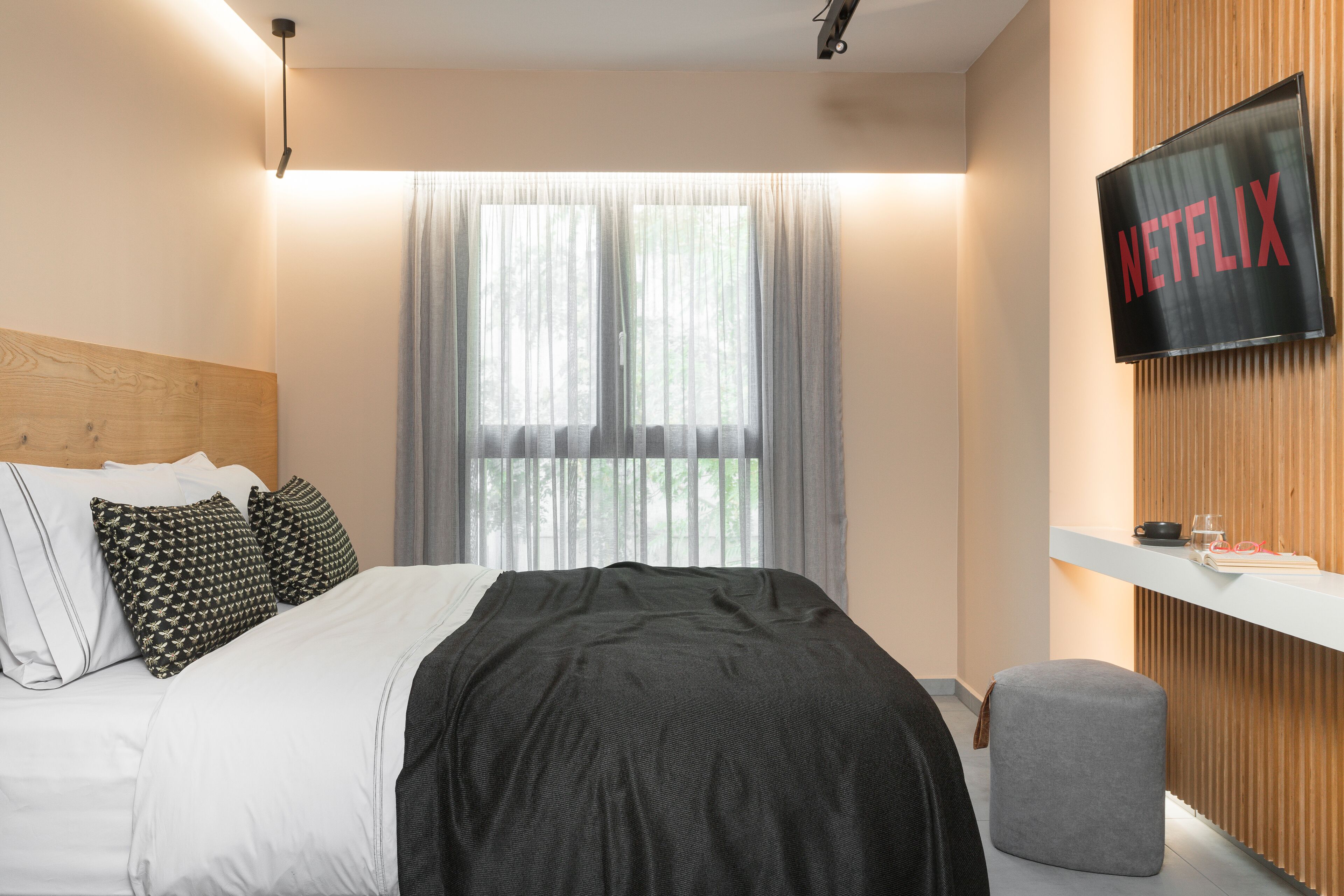 A modern hotel room with a bed, a TV mounted on the wall, and a window with sheer curtains.