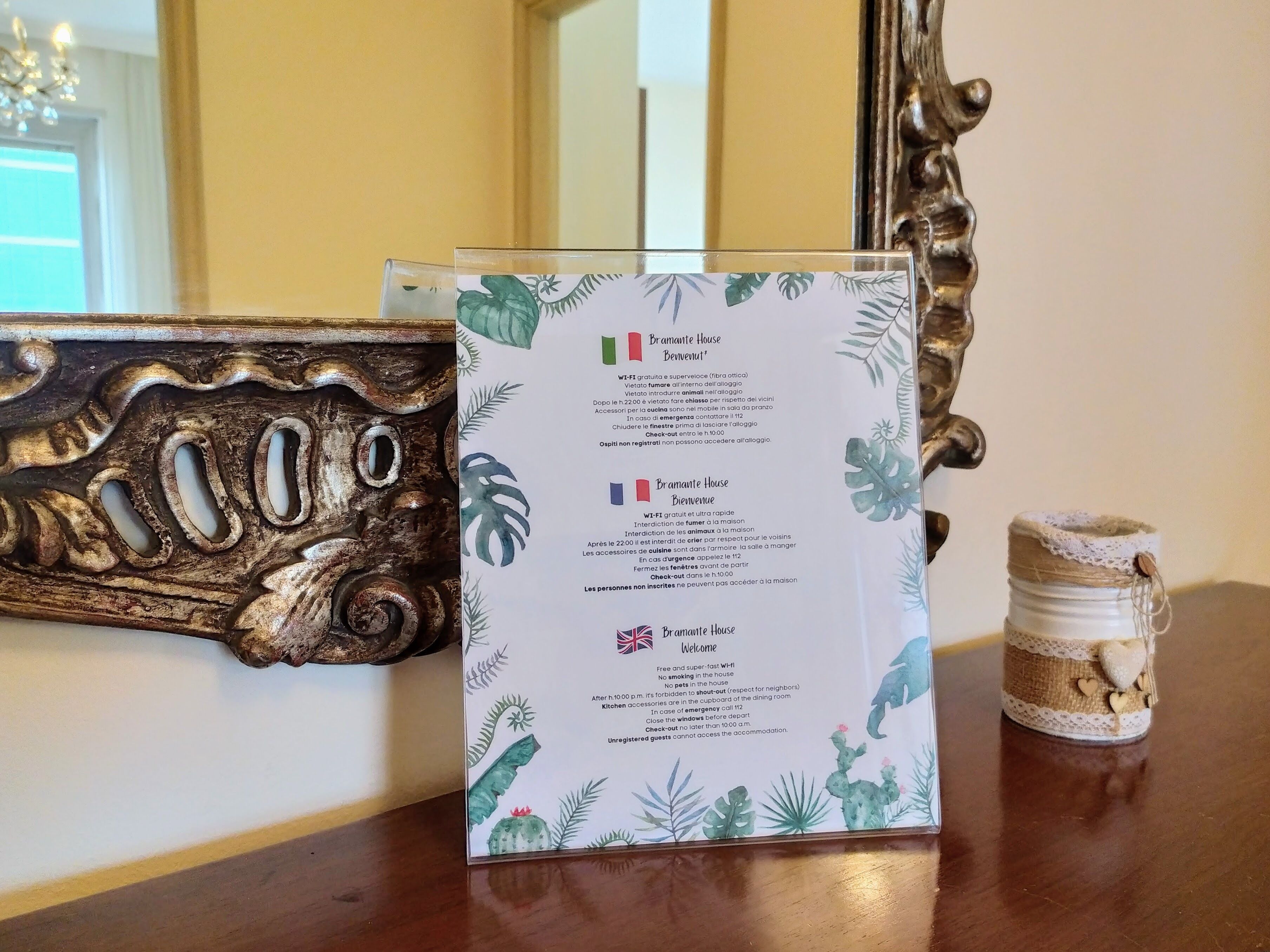 Un cartello di benvenuto con indicazioni in diverse lingue, una cornice decorativa in legno e un piccolo barattolo con un coprilizio in pizzo.