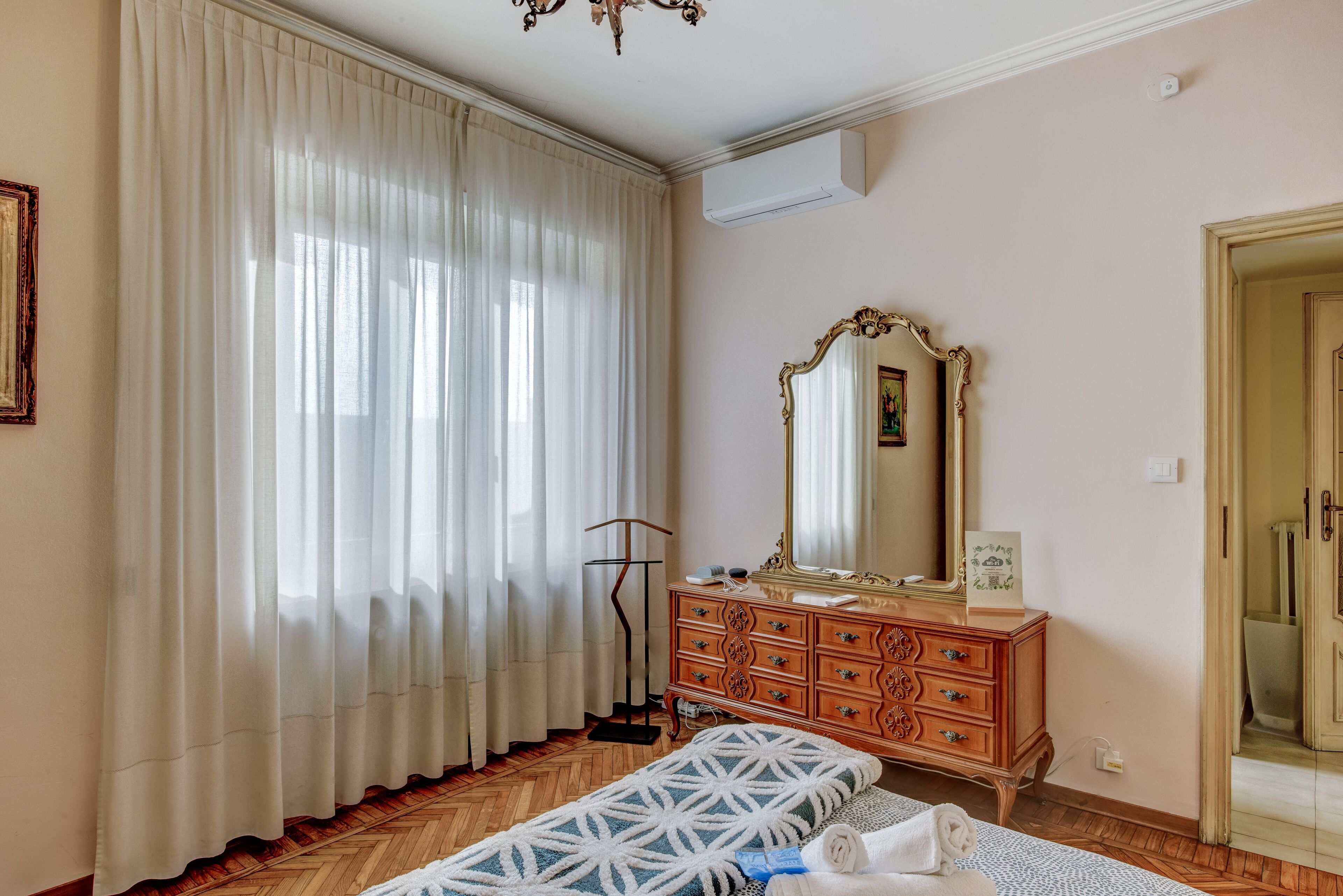 A bedroom with a wooden floor, a bed with a patterned bedspread, a dresser with a mirror, and a window with sheer curtains.