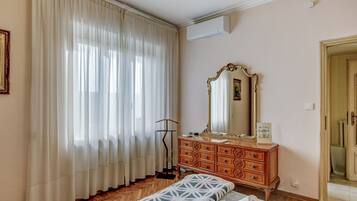 A bedroom with a wooden floor, a bed with a patterned bedspread, a dresser with a mirror, and a window with sheer curtains.
