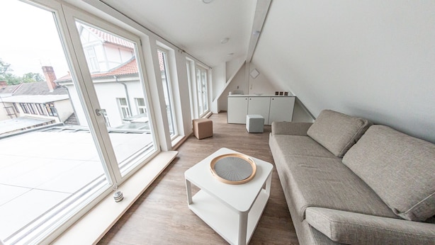 A modern loft apartment with a large window, a grey sofa, a small white table, and a kitchen area. - Harz cosiness (Quedlinburg)