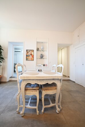 Uma área de refeições com mesa e cadeiras brancas, um quadro na parede e uma estante com objetos decorativos.