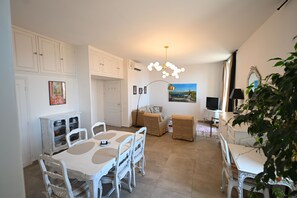 Uma área de refeições com uma mesa e cadeiras brancas, um sofá e uma TV ao fundo.