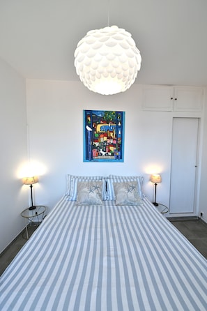 Quarto com colcha listrada, duas luminárias de cabeceira, uma luminária pendente branca grande e uma pintura vibrante de paisagem urbana na parede.