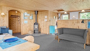 Indretning i en hyggelig hytte med træloft, pejs, sofa, spisebord og et lille køkken.