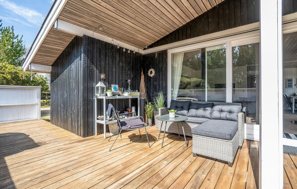 A wooden deck with a wicker sofa and a small table, a white fence, and a large window. - Cozy home in Blåvand with sauna (Blåvand)