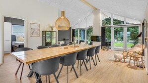 En rummelig spisesal med et langt træbord og moderne stole, store vinduer der giver udsigt over det fri, og en moderne pendellampe.