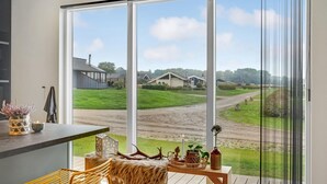 Et moderne køkken med en stor skydedør, der fører ud til en terrasse. Terrassen har udsigt over et landligt landskab med en grusvej og huse.