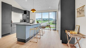 Et moderne køkken med en central ø, et spiseområde med barstole og et stort vindue med udsigt over landskabet.