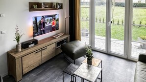 Un salon moderne avec un téléviseur à écran plat fixé au mur, une console en bois, une table basse en verre et un pouf en cuir noir.