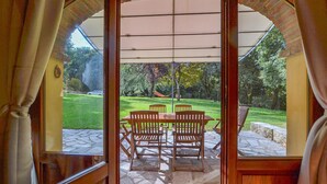 Un patio exterior con una mesa y sillas, un área de estar techada y vistas a un jardín con árboles.