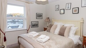 A neatly made bed with a view of a harbor through a window, framed pictures on the wall, and a lamp on the bedside table. - 13 Above Town (Dartmouth)