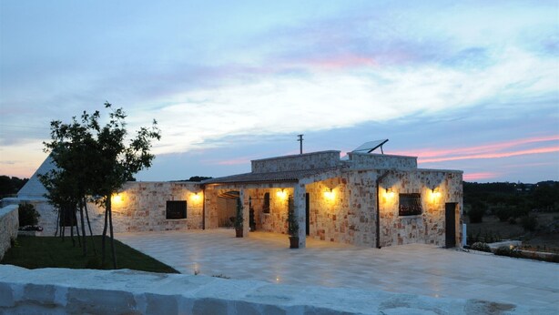 A stone building with multiple rooms and a covered outdoor area, surrounded by a stone wall and a small tree.