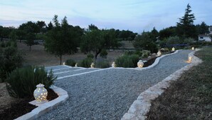 Un chemin de gravier agrémenté de luminaires décoratifs et de plantes, entouré d’arbres et d’un mur de pierres.