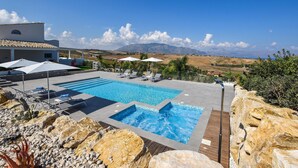 A pool area with lounge chairs and umbrellas, surrounded by rocks and plants, with a view of distant mountains. - Cozy home in Partinico (Partinico)