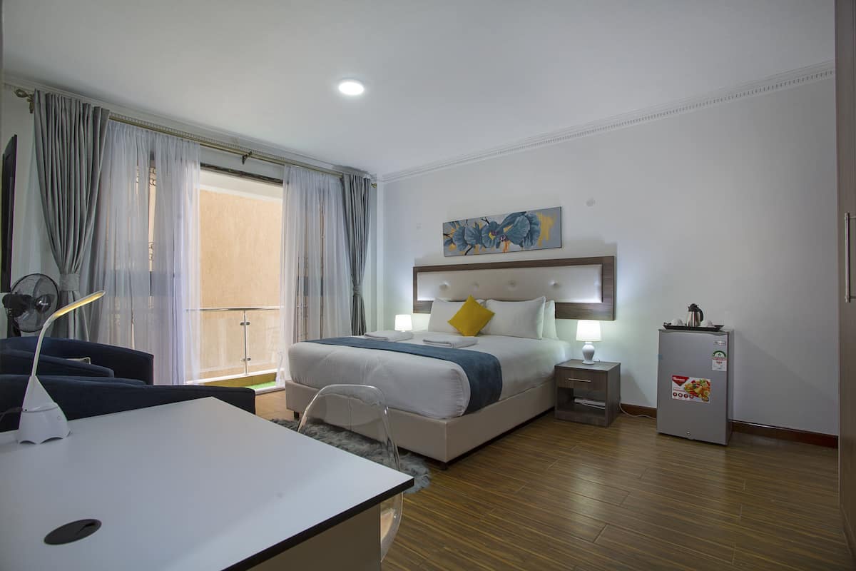 A hotel room with a bed, bedside table, lamp, fan, and a refrigerator.