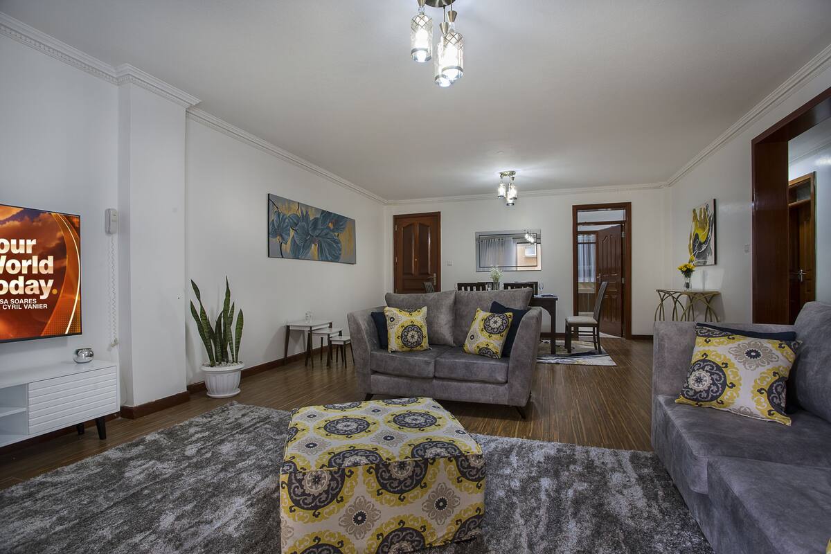 A living room with a grey sofa, a patterned ottoman, a flat-screen TV, and a dining area with a table and chairs.