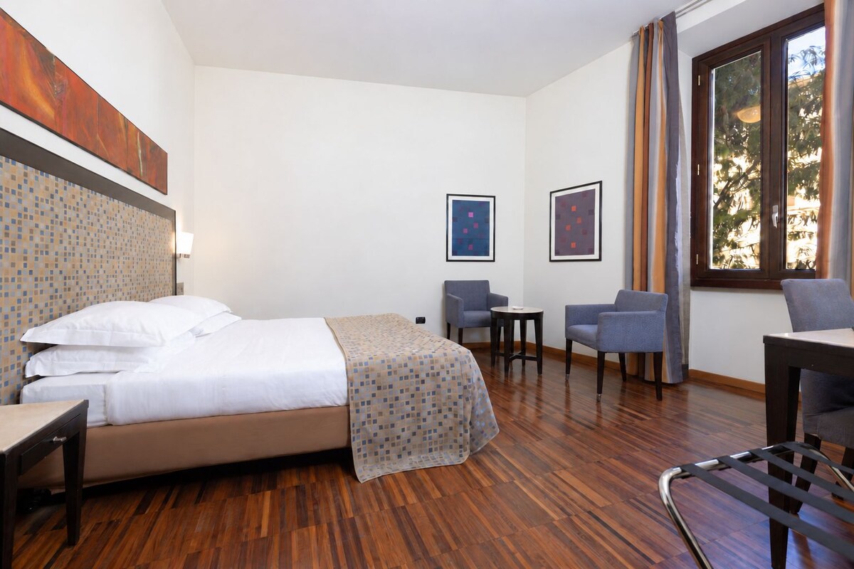 Habitación de hotel con cama, dos sillones, una mesita y obras de arte en las paredes.