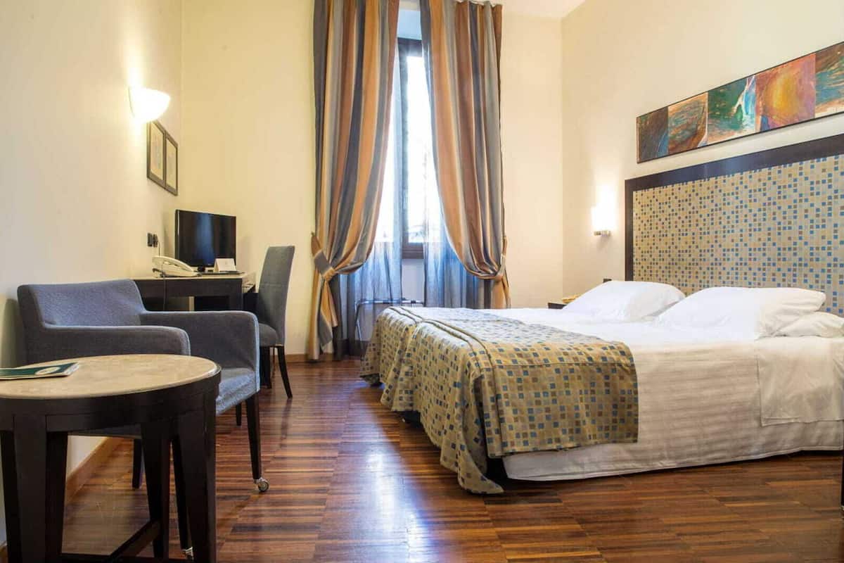 Quarto de hotel com cama, duas cadeiras, uma escrivaninha com computador e um quadro na parede.