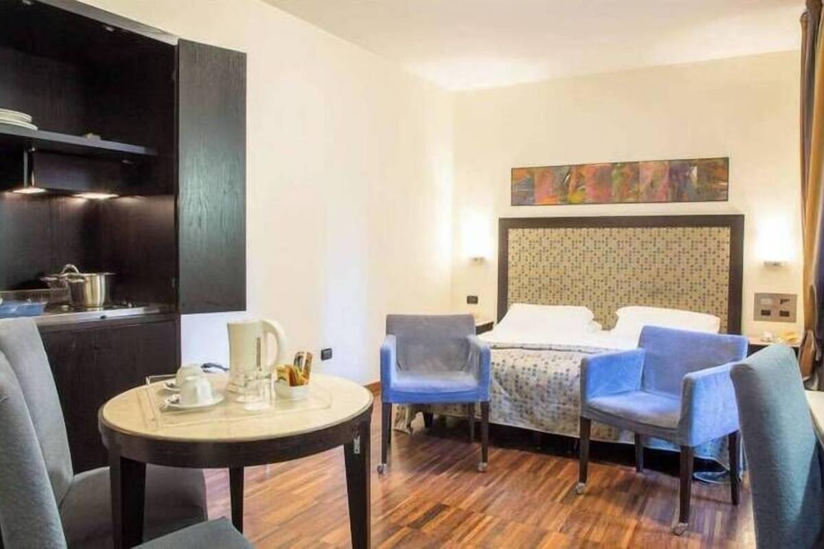 Quarto de hotel com cama, duas cadeiras, uma mesinha com chaleira, um quadro na parede e uma área de cozinha compacta.