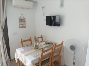 A dining area with a table set for four, a television mounted on the wall, and a standing fan.