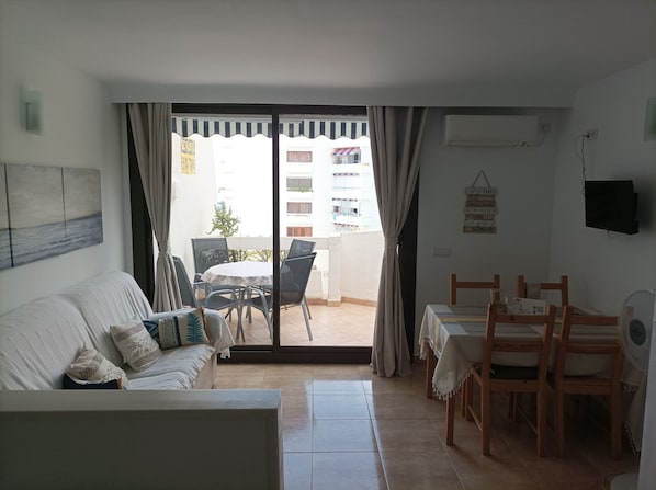 A living room with a sofa, a dining table with chairs, and a sliding glass door leading to an outdoor area with a table and chairs.