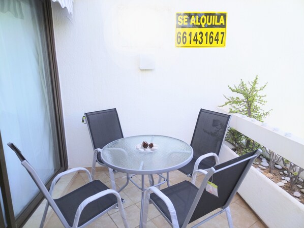 A small outdoor balcony with a glass table and four chairs, a sign with a phone number, and a view of a garden with small trees and rocks.