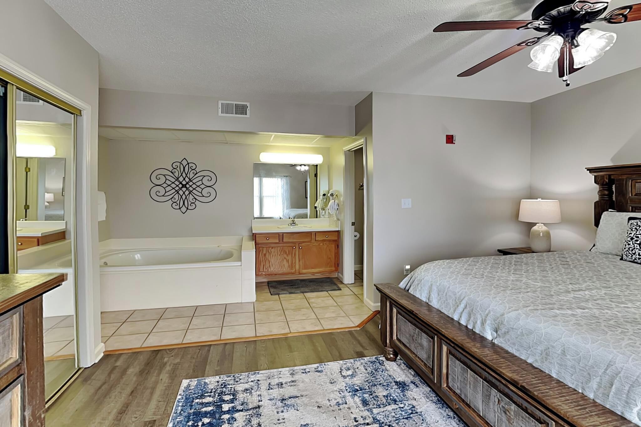 A bedroom with a bed, a nightstand, a lamp, a ceiling fan, a bathroom with a bathtub, a sink, and a mirror.