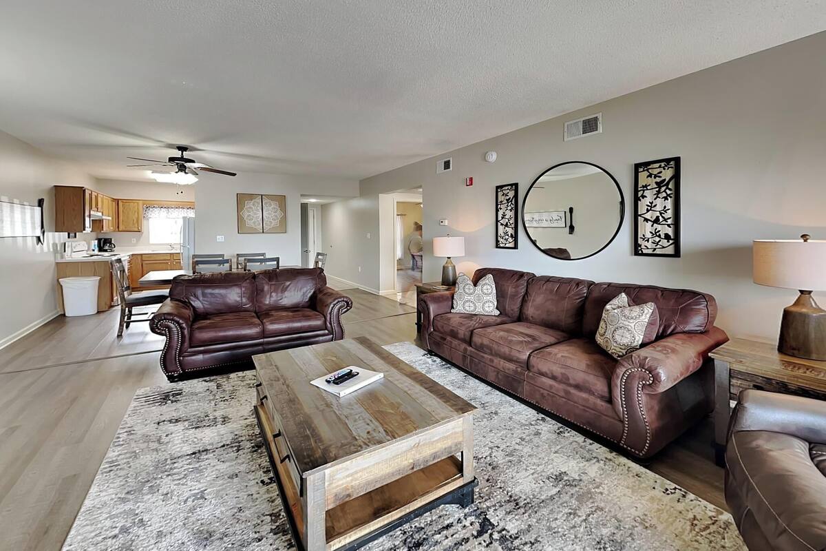 A spacious living room with two brown leather sofas, a wooden coffee table, and a kitchen in the background.