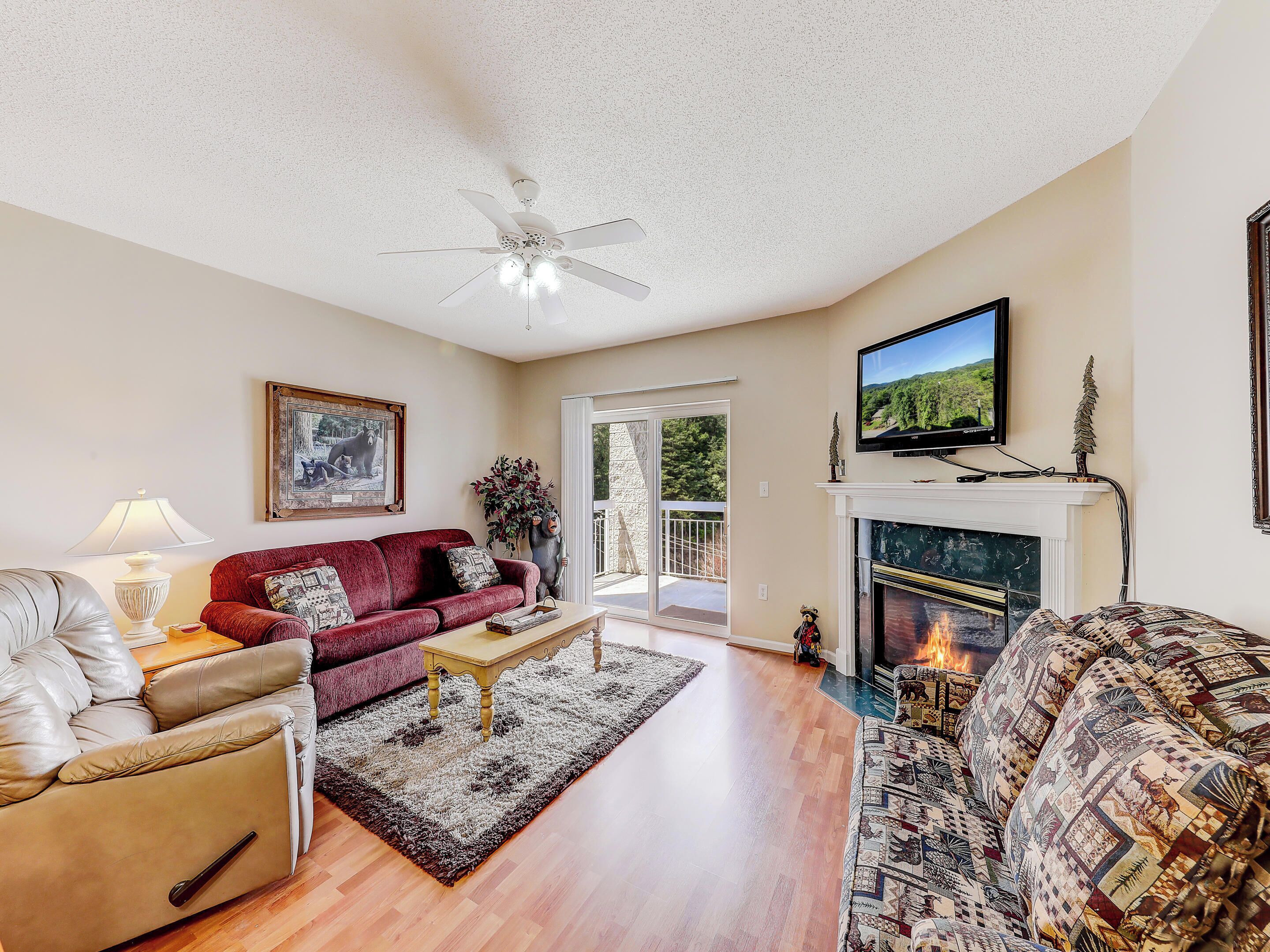A spacious living room with a fireplace, a ceiling fan, a television, and a large window.