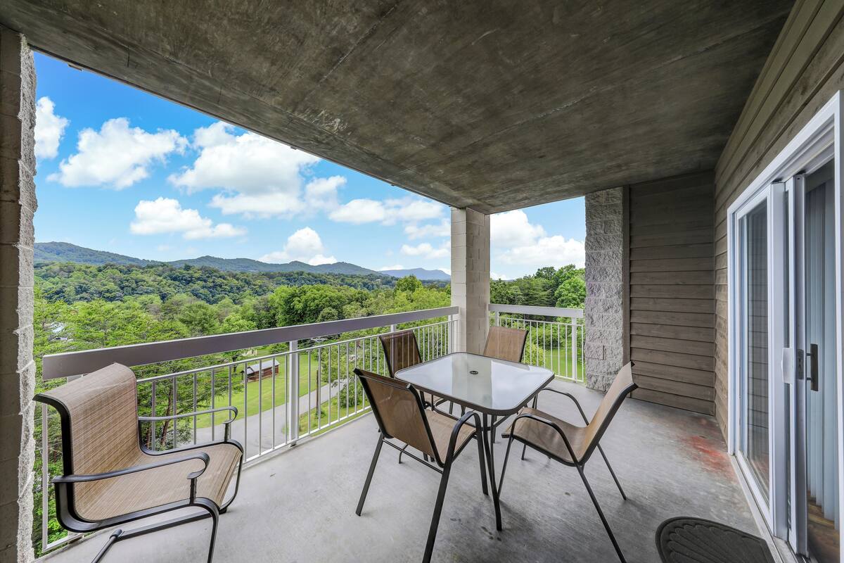 A balcony with a table and chairs, a view of green hills and mountains, and a concrete ceiling.
