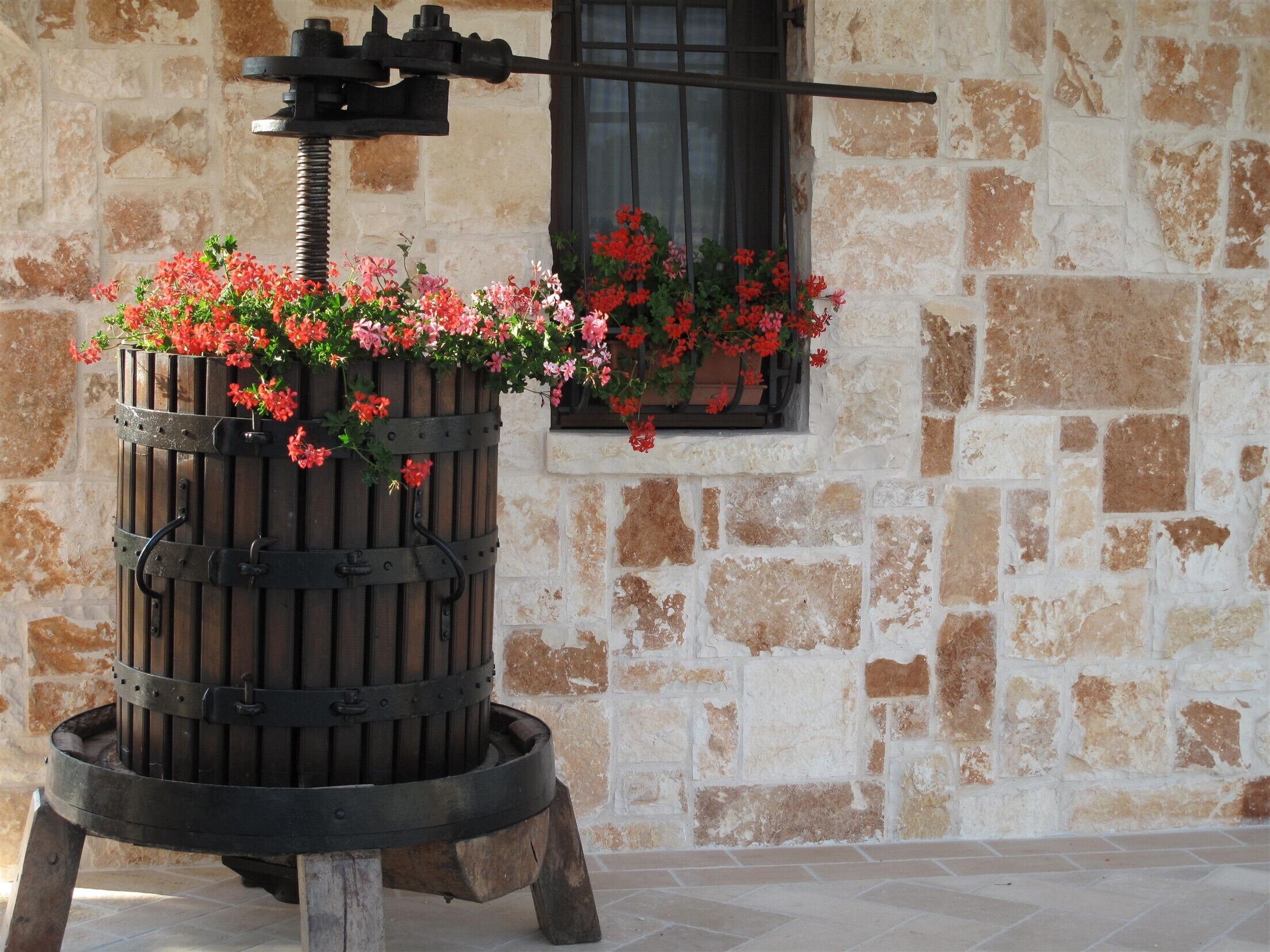Una bomba de agua rústica al aire libre con una maceta de flores unida, frente a un muro de piedra con una ventana.