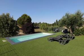 Une piscine rectangulaire bordée d’un liseré foncé, entourée de gazon artificiel. On y trouve deux chaises longues noires au design original, ainsi qu’un petit espace jardin avec un banc et un arbre.