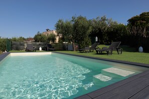 Una piscina rectangular con agua cristalina, rodeada por una terraza de madera y una zona verde con tumbonas.