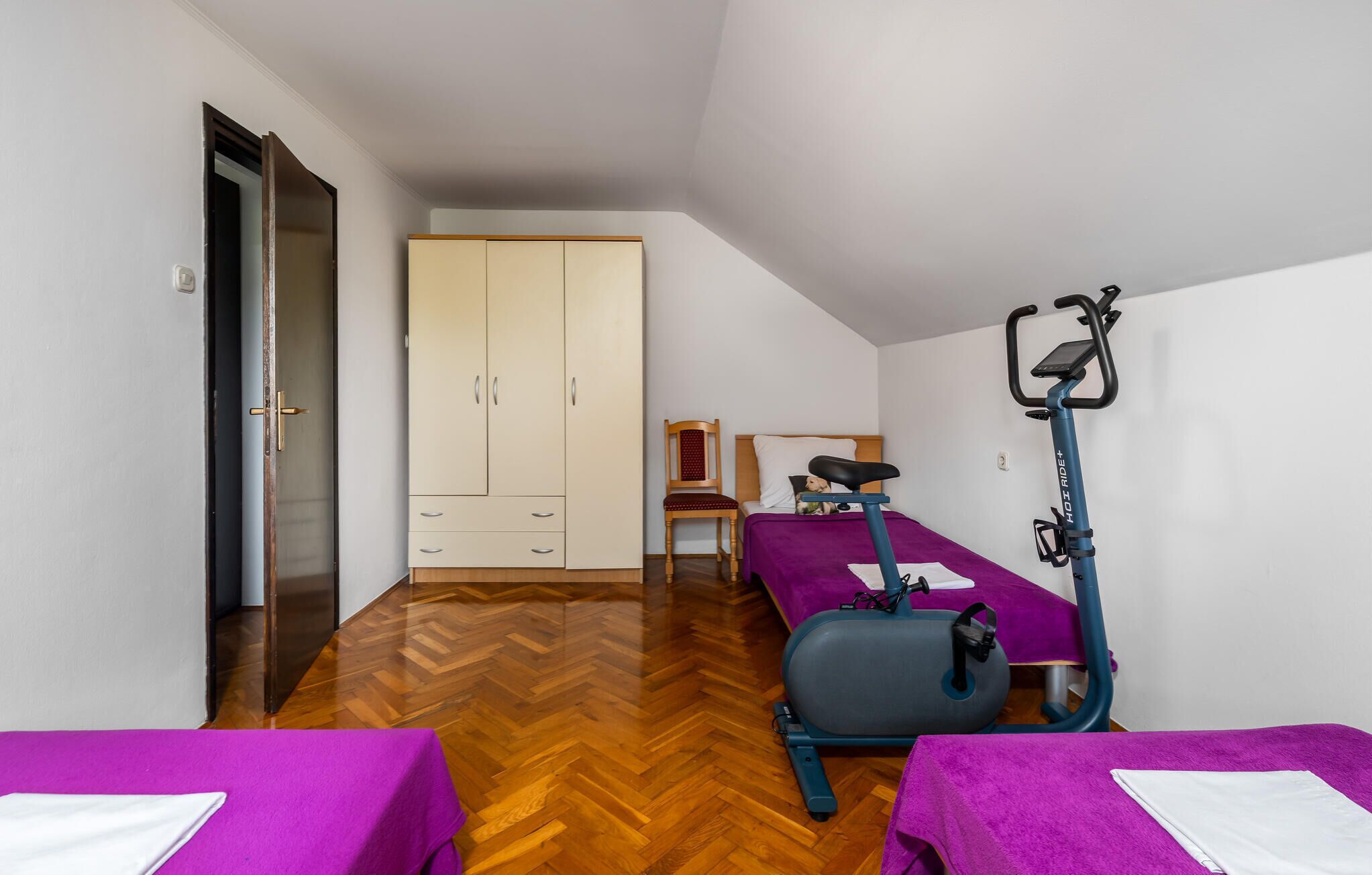Habitación de hotel con suelo de parqué tipo espiga, dos camas con ropa de cama morada, un armario, una silla y una bicicleta estática.