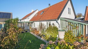 Une maison blanche avec un toit recouvert de tuiles rouges, une véranda et un jardin bien entretenu où poussent différentes plantes et fleurs.