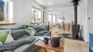 Un salon chaleureux avec un canapé, une table sur laquelle se trouvent une bouilloire et une tasse, une cheminée, et un coin repas à l’arrière-plan.