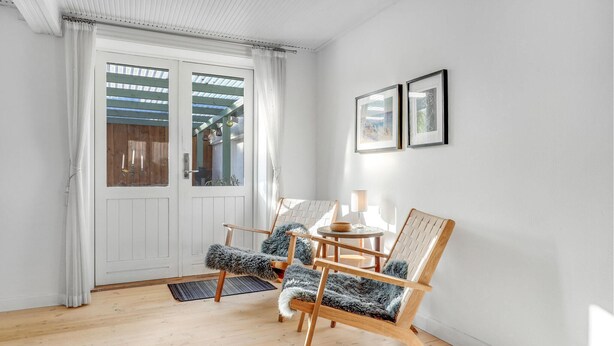 Une pièce avec un plancher en bois, des murs blancs, une porte blanche avec des panneaux de verre, deux fauteuils ornés de coussins en fausse fourrure, une petite table ronde avec une lampe et deux tableaux encadrés accrochés au mur.