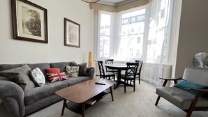 Un salon avec un canapé gris, une table basse en bois, une table à manger avec des chaises et une fenêtre avec des rideaux.