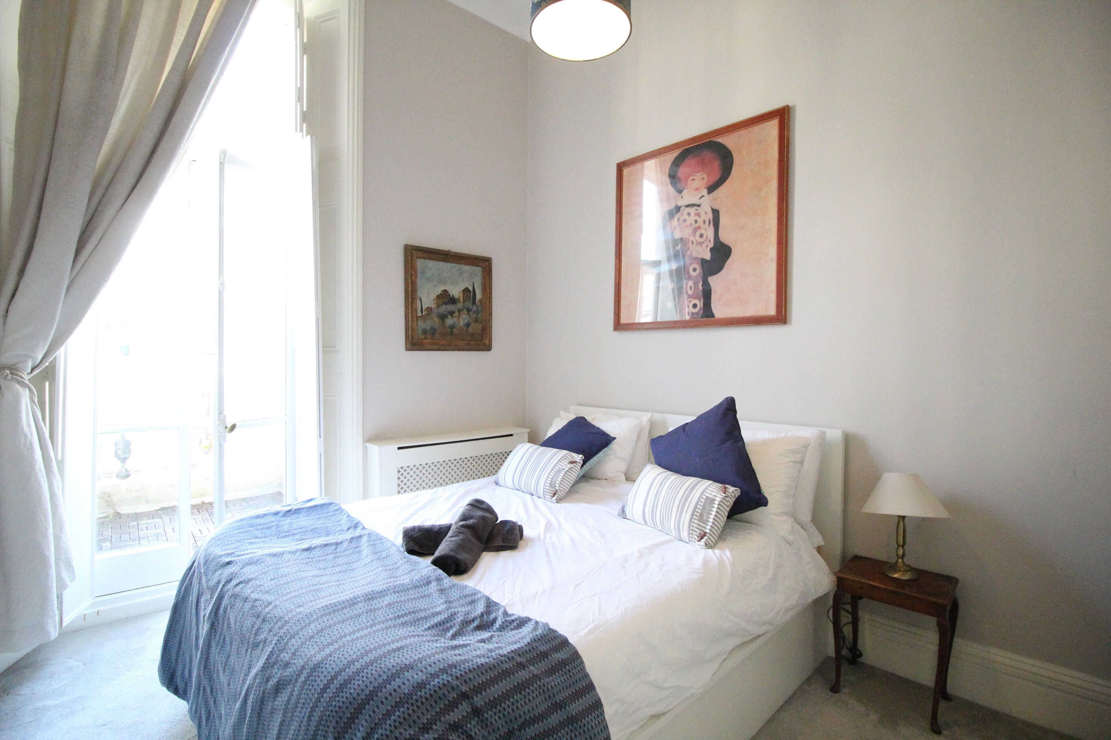 A bedroom with a large bed, a bedside table with a lamp, a window with curtains, and two framed pictures on the wall.