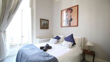 A bedroom with a large bed, a bedside table with a lamp, a window with curtains, and two framed pictures on the wall.