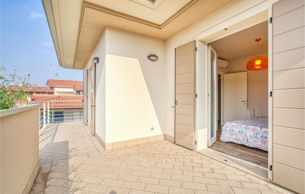 A balcony with a tiled floor, a door leading to a bedroom with a bed and a patterned bedspread, and a view of neighboring buildings.
