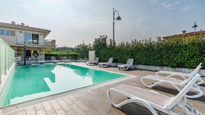 Una piscina con sillas de descanso, un edificio con balcones y un seto al fondo.