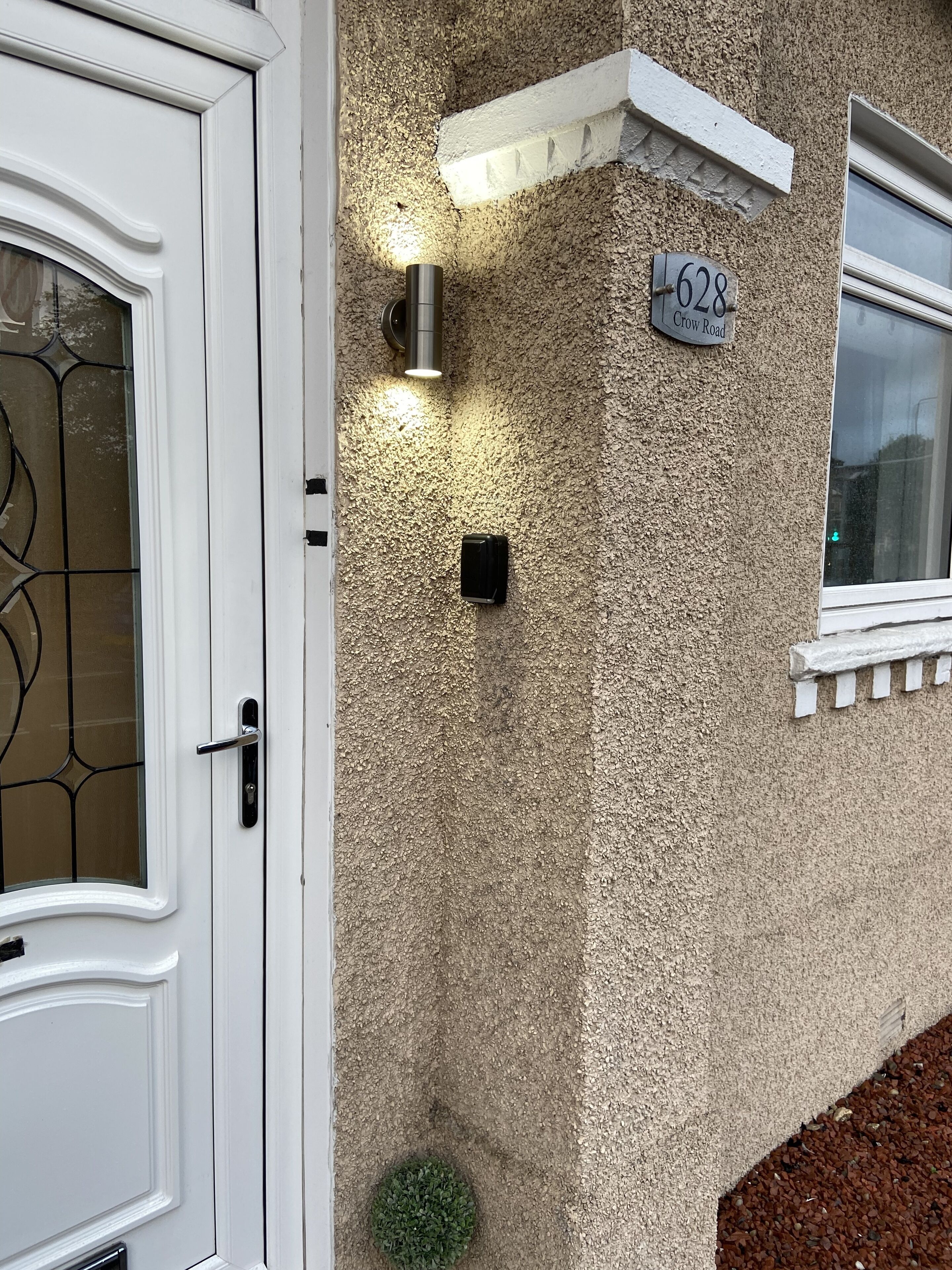 Een huis met een witte deur, een raam met een witte omlijsting, een wandlamp, een kleine potplant en een huisnummerbord.
