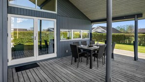 En overdækket terrasse med bord og stole, en skydedør samt udsigt til en have og nabohuse.