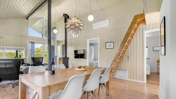 Et moderne spiseområde med et træbord, hvide stole, en stor hængende lampe og en trappe til et andet rum.
