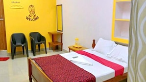 Une chambre d’hôtel avec un lit en bois, une couvre-lit rouge et blanche, deux chaises noires, une petite table en bois et un mur jaune orné d’une pancarte « BIENVENUE ».