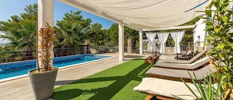 A poolside area with artificial grass, lounge chairs, and a pergola with white fabric.